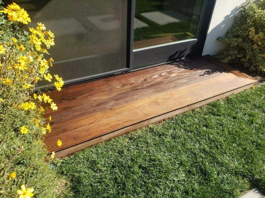 A wooden deck leading to a glass door, surrounded by vibrant yellow flowers and lush green grass under bright sunlight.