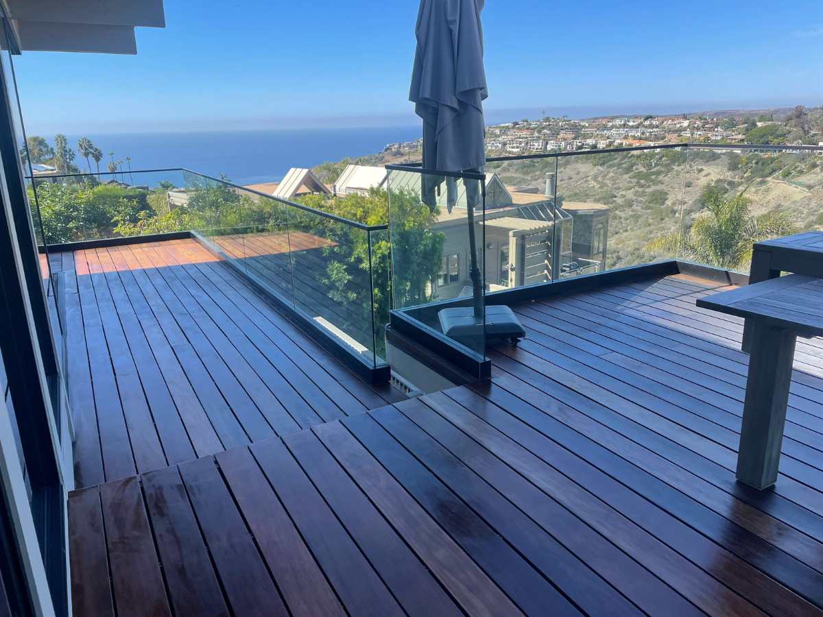 A spacious wooden deck with glass railing overlooks a coastal view of palm trees and hillside homes under a clear blue sky.