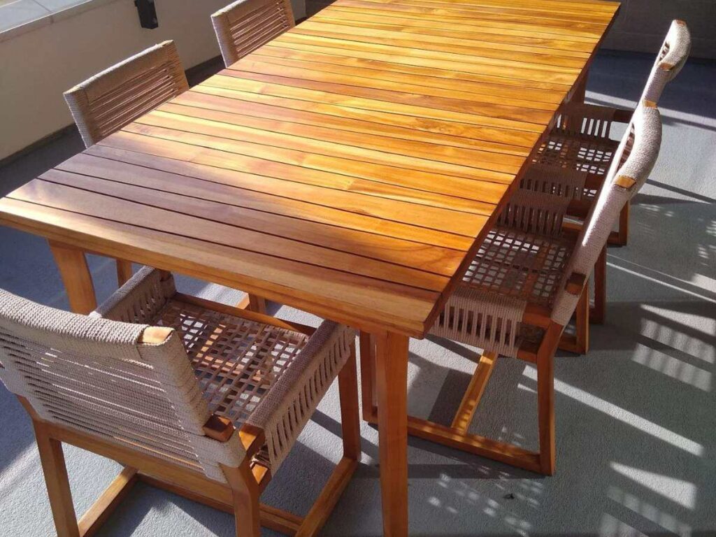 A wooden dining table with a smooth top and four woven chairs, set in a sunny outdoor space, casting elegant shadows.