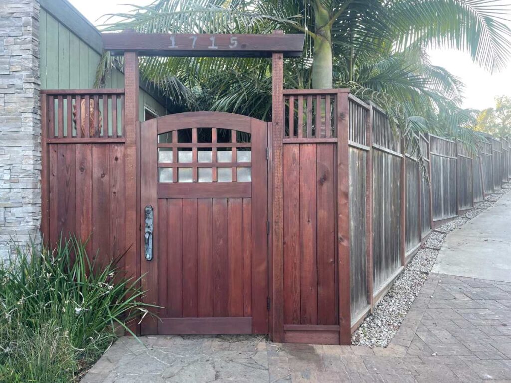 A wooden gate with a rounded top and decorative paneling, flanked by palm trees and a stone wall, marked with the number 1715.