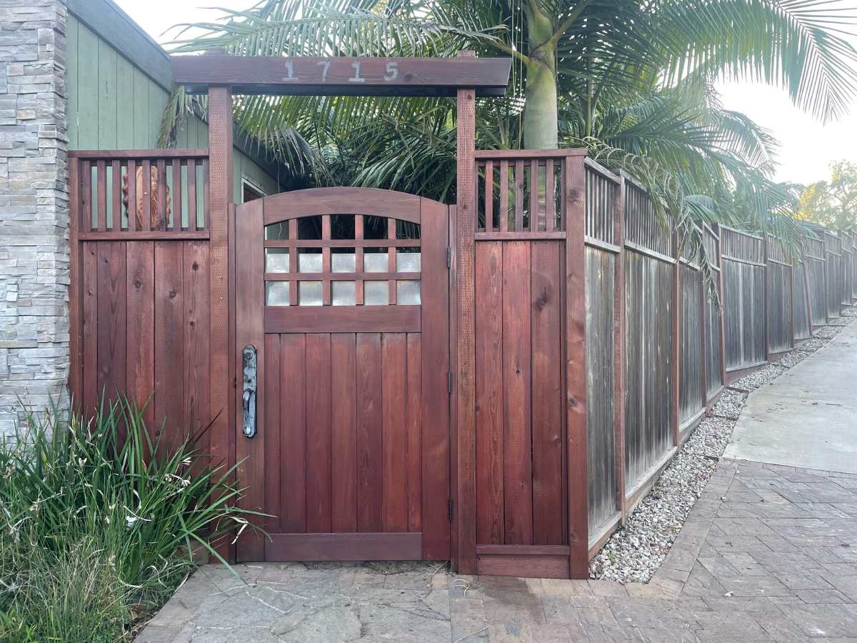 A wooden gate with a rounded top and decorative paneling, flanked by palm trees and a stone wall, marked with the number 1715.