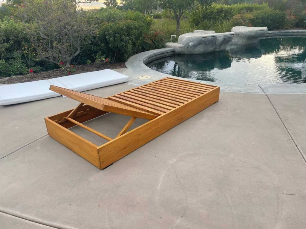 A wooden lounge chair with slatted design positioned beside a pool, surrounded by lush greenery and another white cushion nearby.