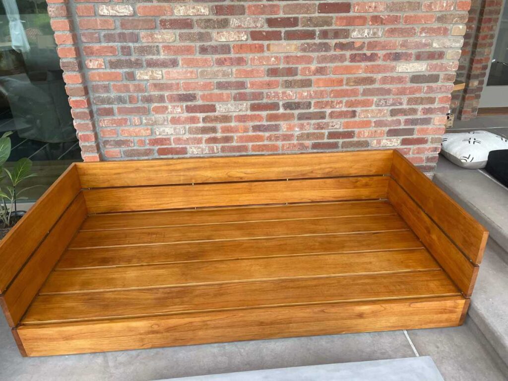 A wooden outdoor bench with a polished finish, set against a textured brick wall, providing a cozy seating area.