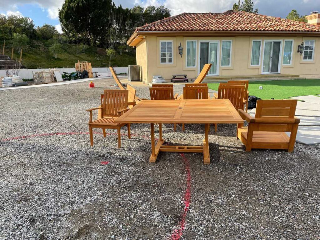 A wooden patio dining set with a table and three chairs is placed on a gravel area, with a house and greenery in the background.
