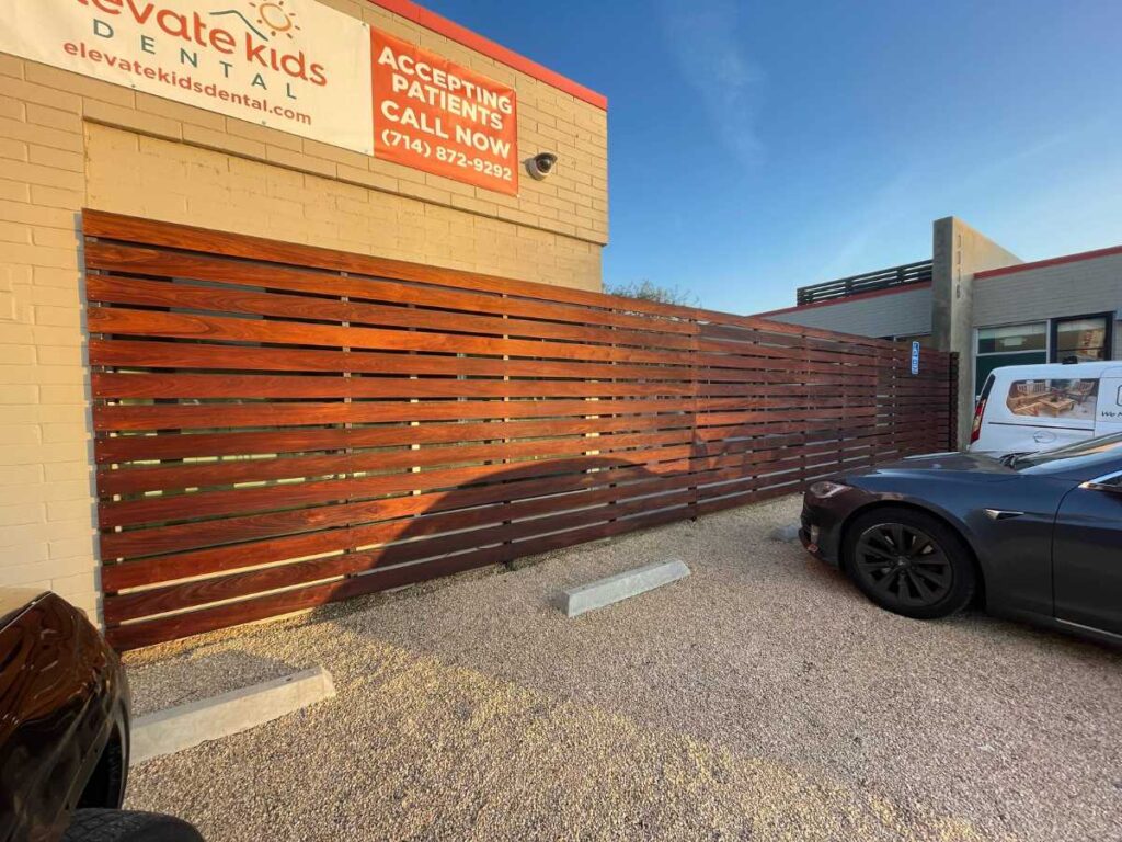A wooden slat fence beside a dental office, with a sign announcing patient acceptance and two parked cars in a gravel lot.