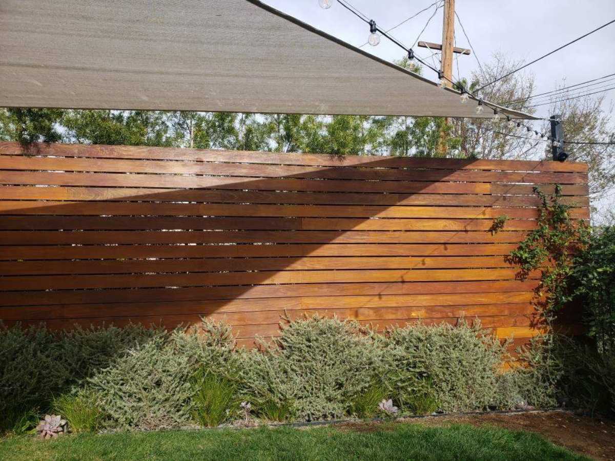 A wooden slat fence with greenery below, shaded by a fabric canopy, and string lights above, in a serene outdoor setting.