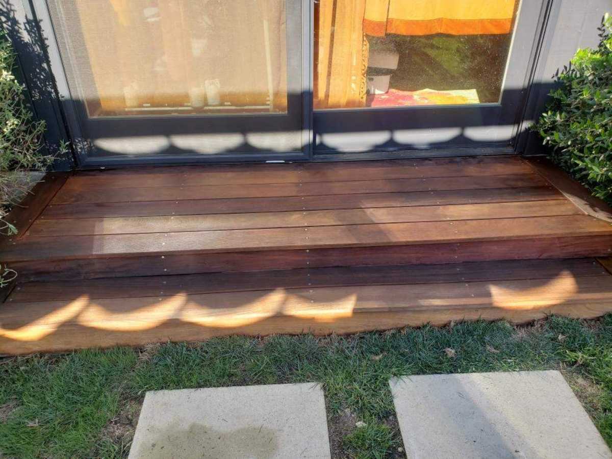 Wooden steps lead to a glass door, surrounded by greenery and sunlight casting decorative shadows on the polished surface.